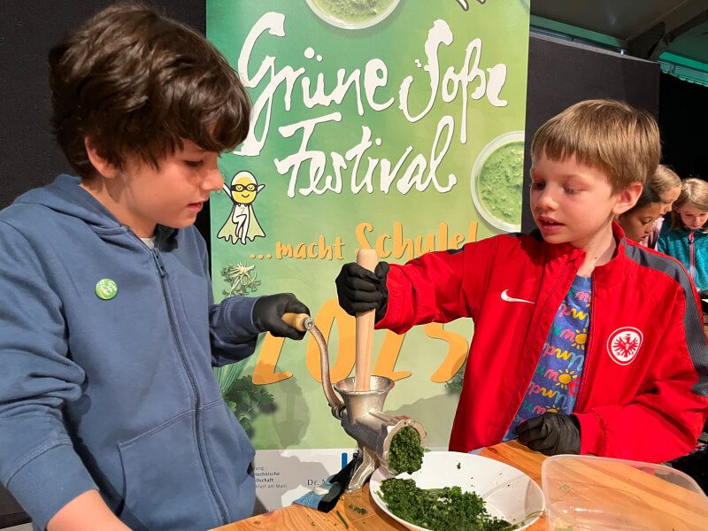 Zwei Grundschulkinder in Handschuhen drehen gemeinsam eine Gemüsequetsche beim Grüne Soße Festival, im Hintergrund ein Plakat mit dem Slogan „macht Schule“.