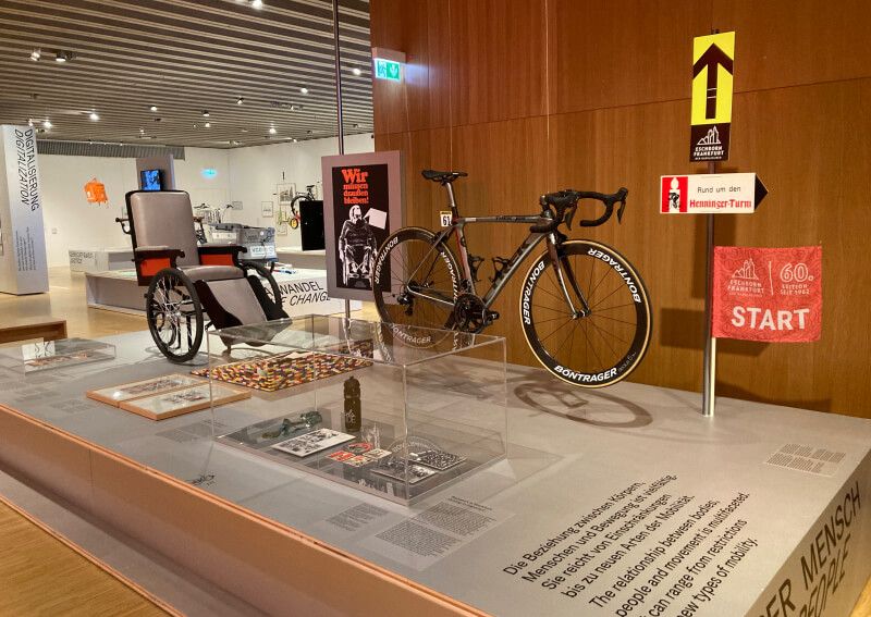 Ausstellungsansicht mit Rennrollstuhl und Rennrad im Museum, ergänzt durch Plakate, historische Dokumente und ein „START“-Schild des Radrennens rund um den Henninger-Turm.
