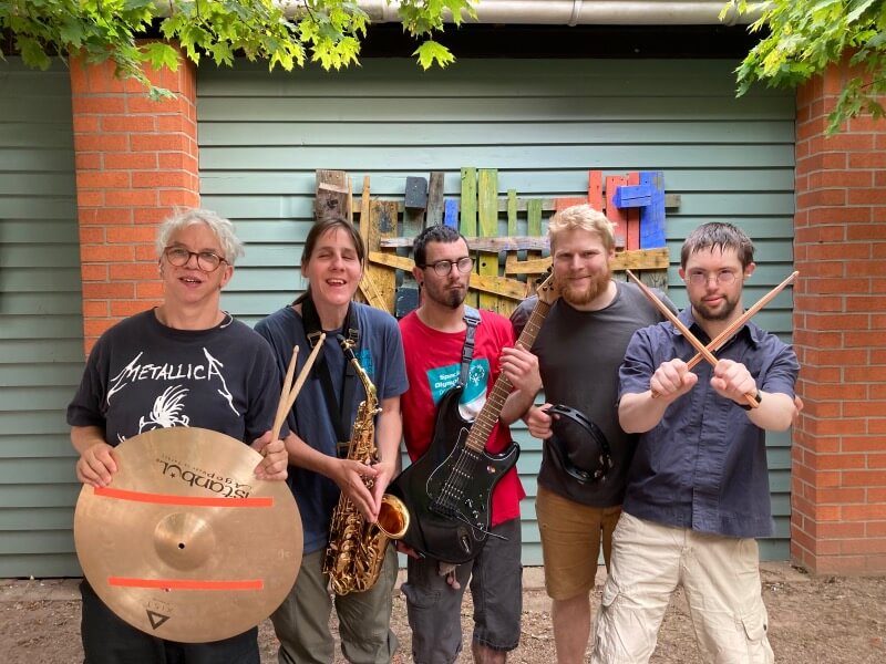 Fünf Musiker mit verschiedenen Instrumenten wie E-Gitarre, Saxofon und Schlagzeug posieren lachend vor einem bunten Wandkunstwerk aus Holz.