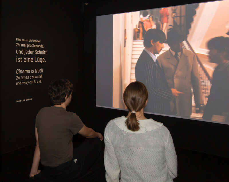 Zwei Personen sitzen in einem abgedunkelten Ausstellungsraum und schauen einen Filmausschnitt; Zitat von Jean-Luc Godard zur Wahrheit des Films an der Wand.