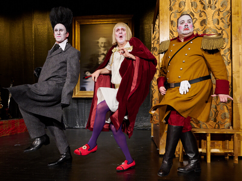 Drei Schauspieler in expressiven historischen Kostümen auf der Theaterbühne während einer humorvollen und stilisierten Szene mit überzogener Mimik.