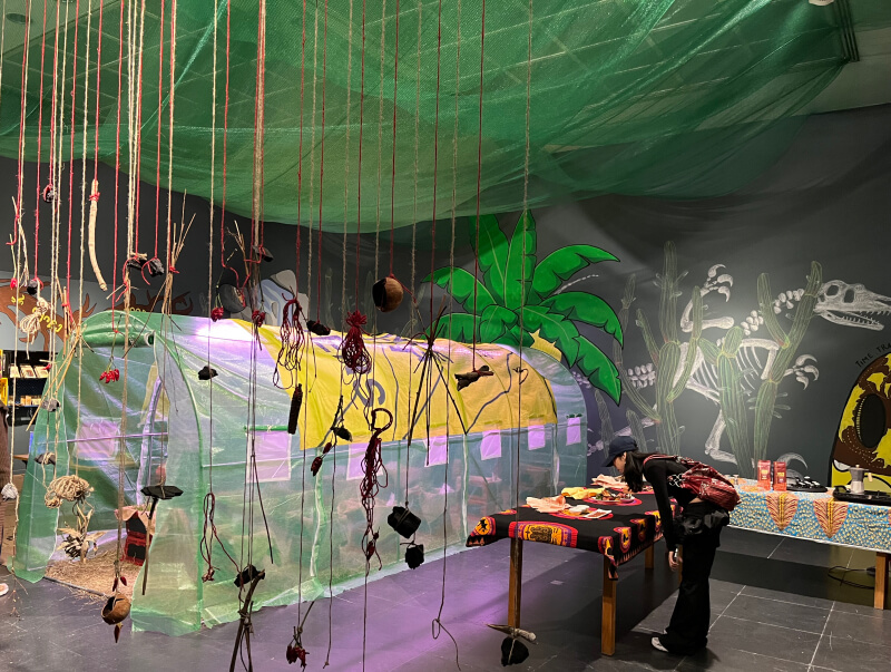 Blick in eine künstlerisch gestaltete Ausstellungsinstallation mit Gewächshaus, bunten Textilien und aufgehängten Naturmaterialien im Vordergrund.