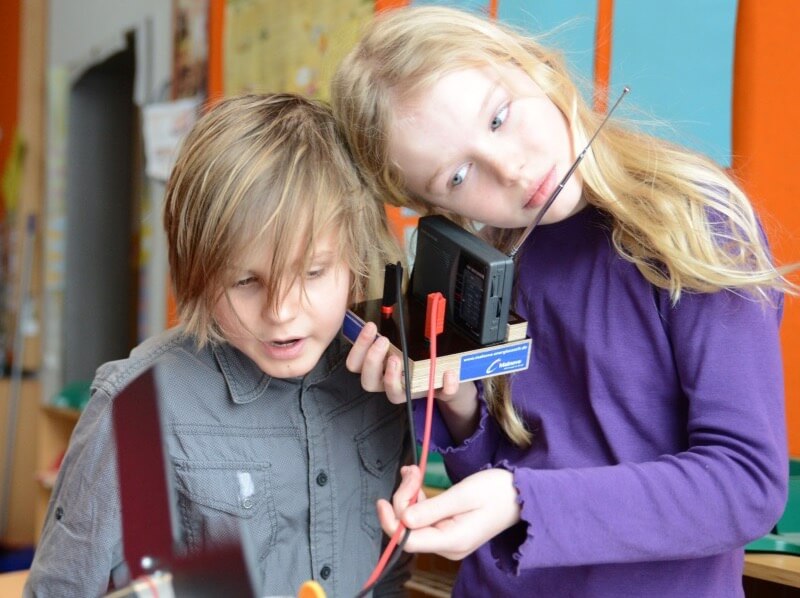 Zwei Grundschulkinder experimentieren mit einem selbstgebauten Radio oder technischen Gerät im Klassenzimmer und hören gespannt über Antenne und Kopfhörer.